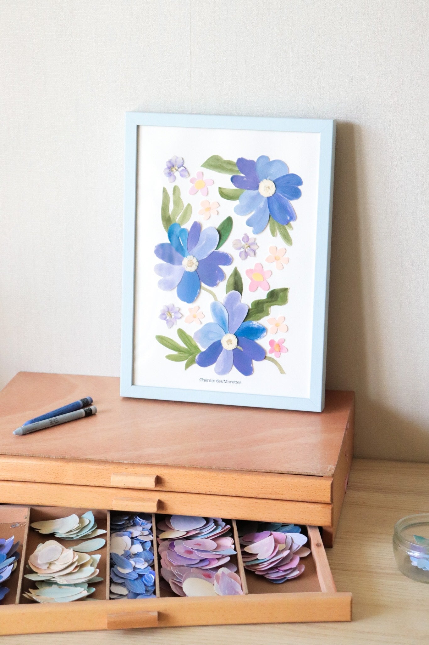 herbier original A4 nommé céleste, collage de fleurs en papier en relief et fleurs pressées sur fond blanc, encadré dans un cadre en bois bleu, création Chemin des Marettes.