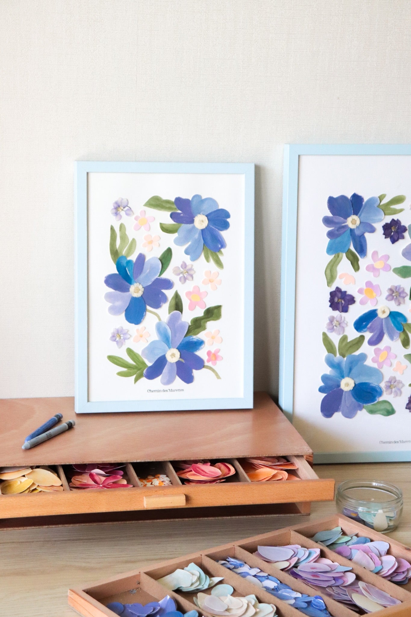herbier original A4 nommé céleste, collage de fleurs en papier en relief et fleurs pressées sur fond blanc, encadré dans un cadre en bois bleu, création Chemin des Marettes.